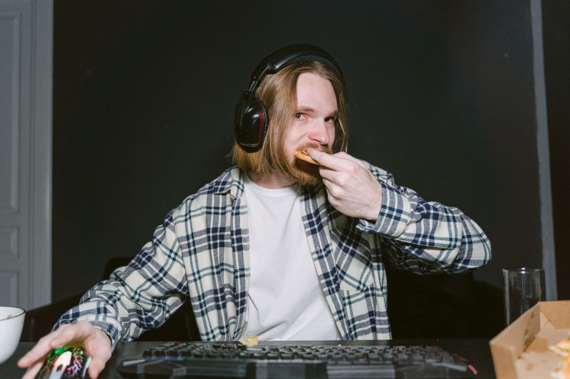 A man wearing headphones eats pizza while gaming on a computer, focused and engaged.