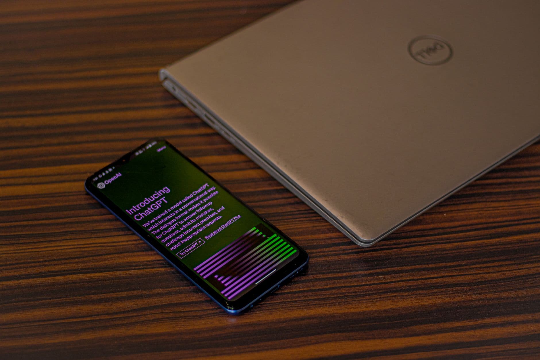 A smartphone displaying ChatGPT app beside a closed laptop on a wooden table.