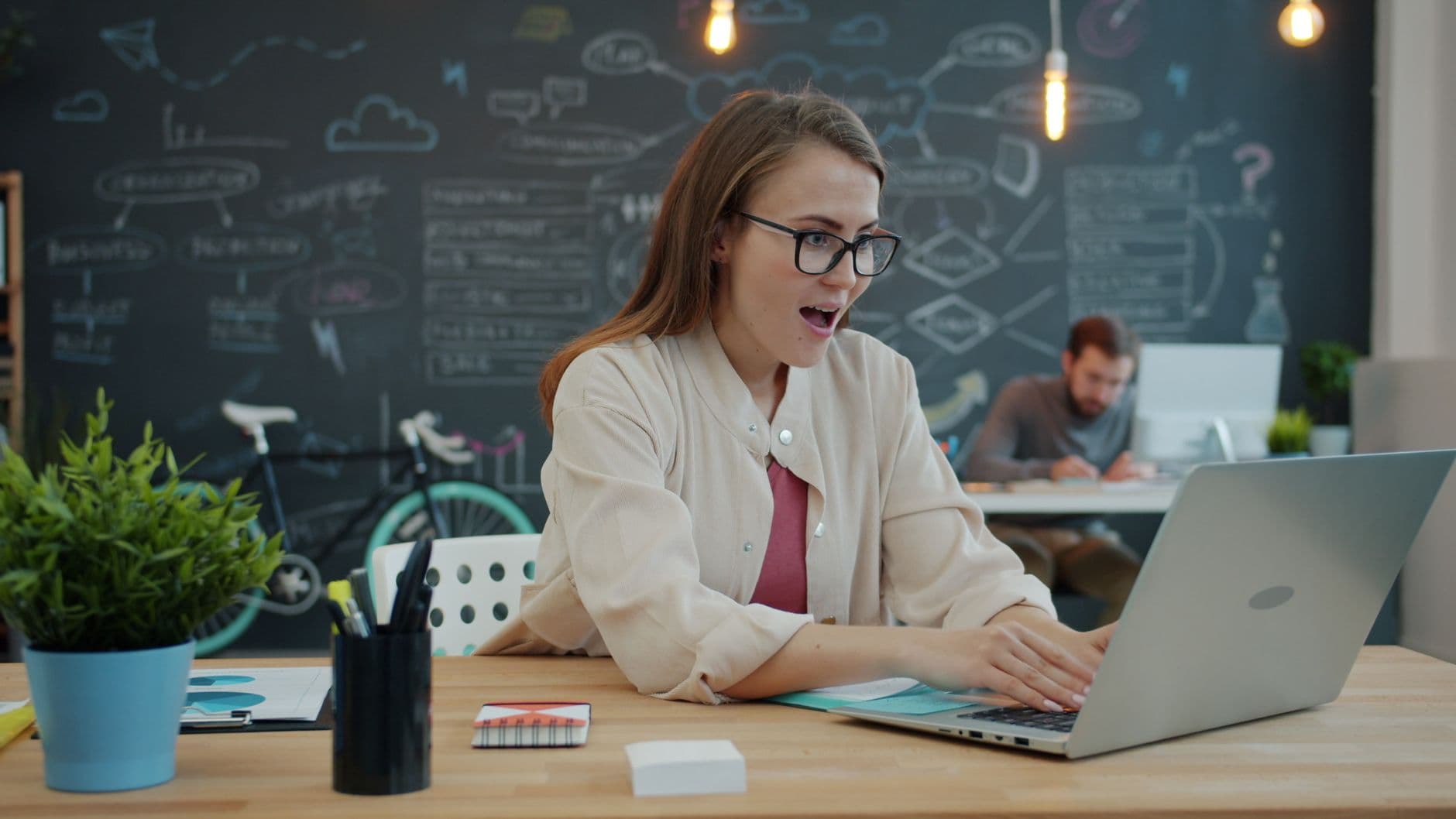 Smiling woman in office amazed by laptop, creative workspace with blackboard in background.