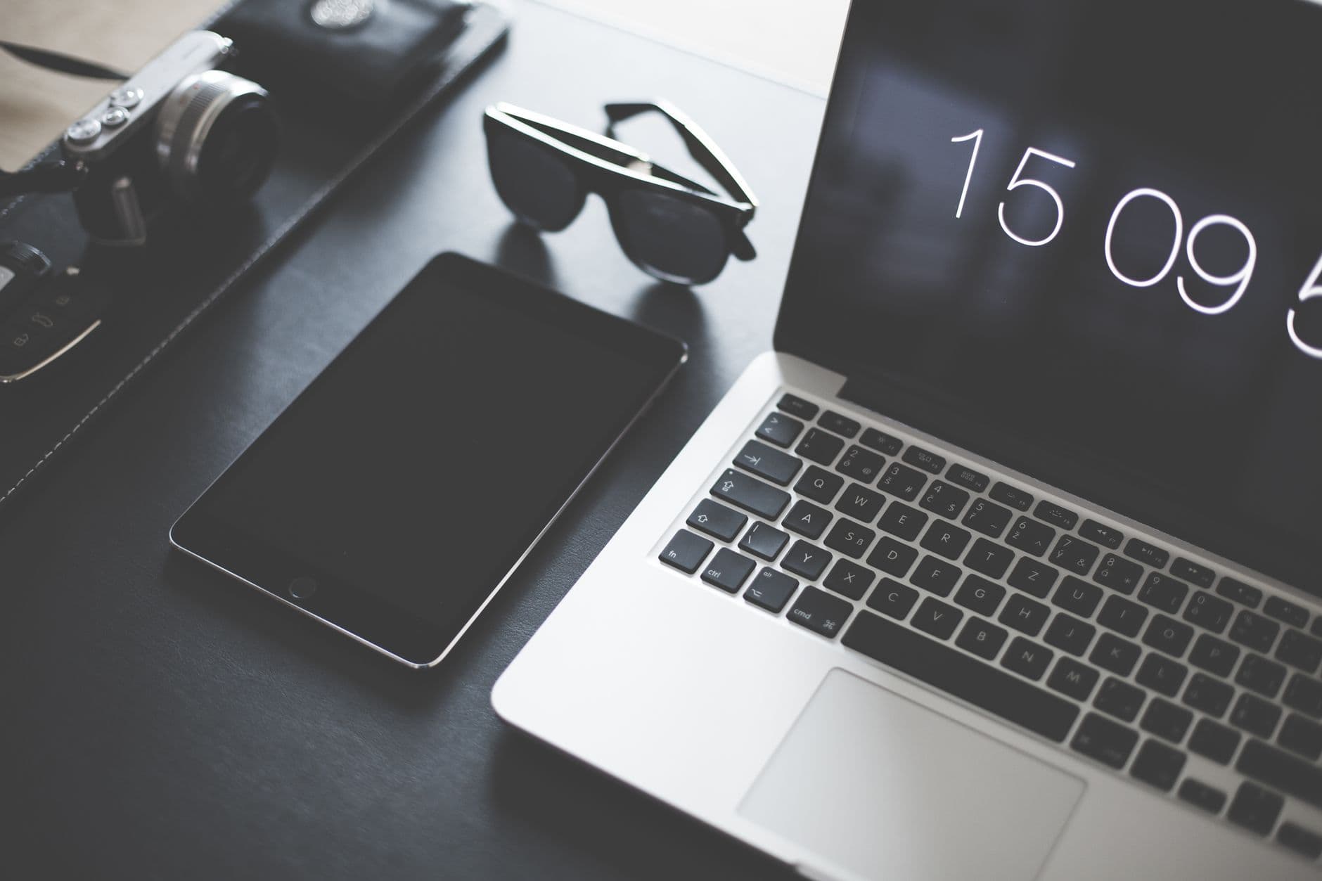 A flat lay of a modern workspace featuring a laptop, tablet, camera, and sunglasses.