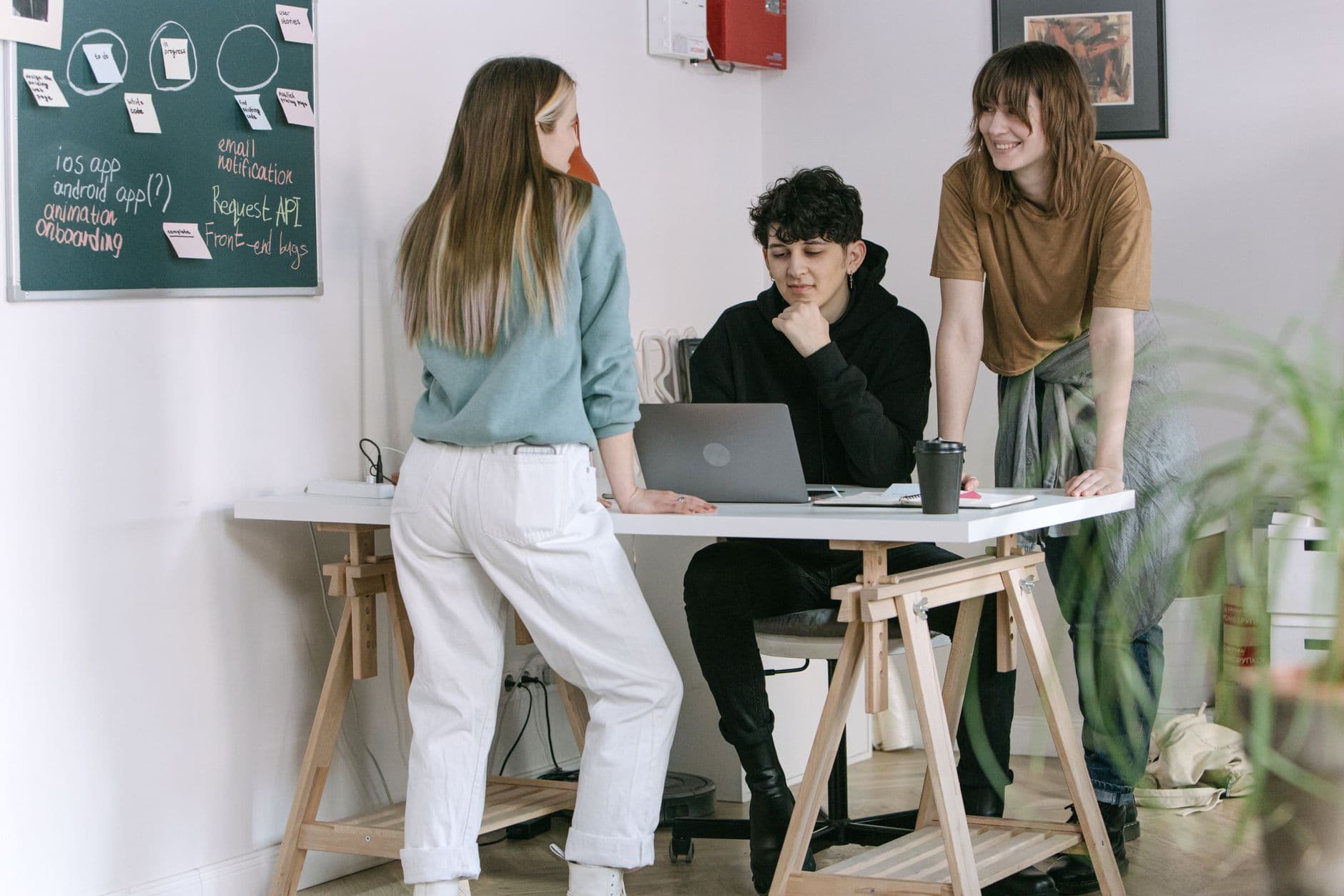 Three young professionals brainstorm ideas around a laptop in a modern office setting.