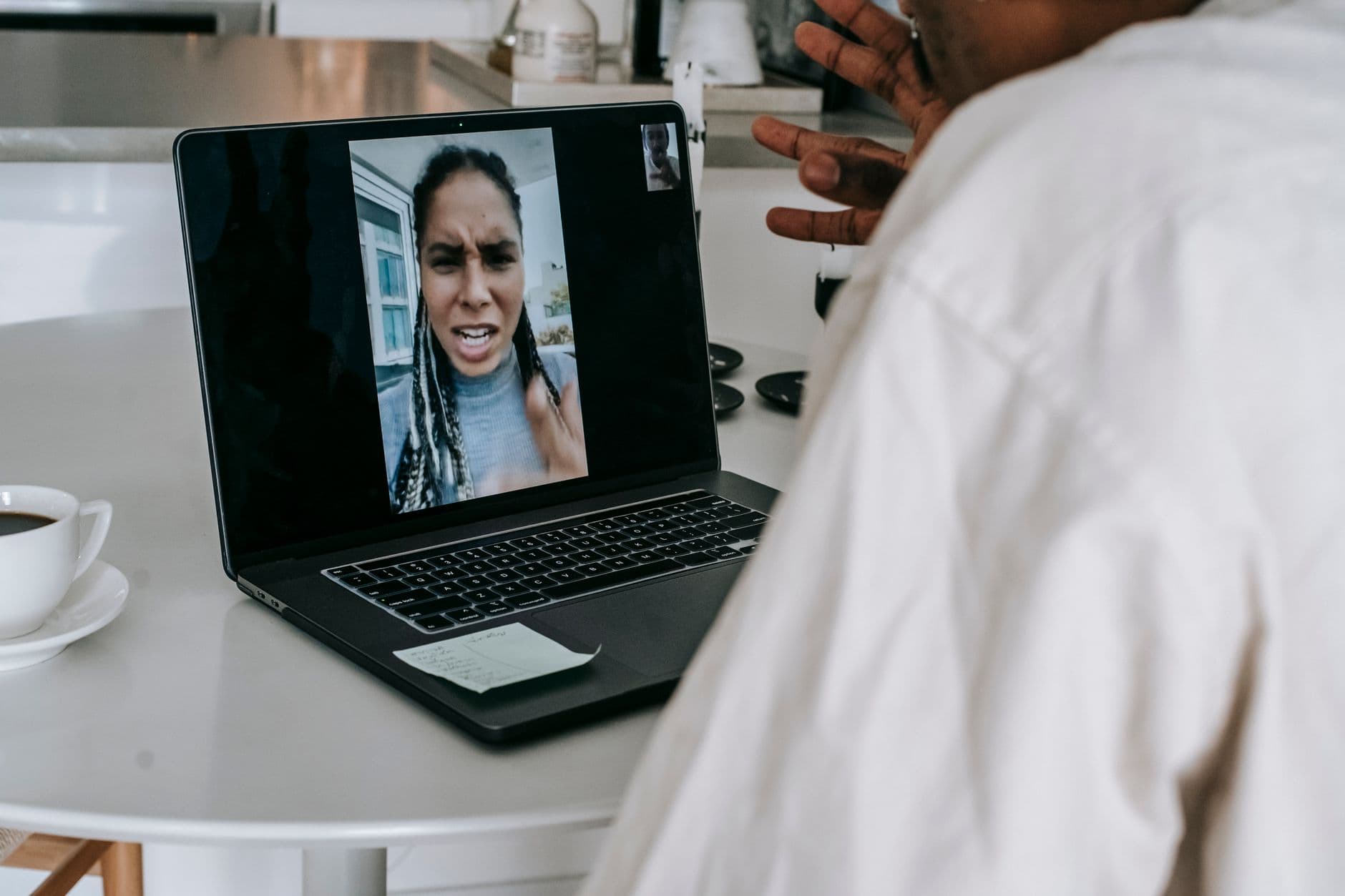 Crop anonymous black male sitting at table in modern kitchen with cup of coffee while having video conversation on laptop and arguing with angry girlfriend